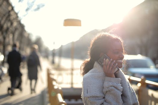 Smiling woman having a phone conversation outdoors with a bright sunny backdrop.