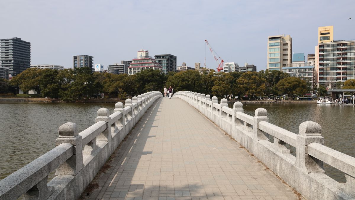 후쿠오카 — A wide stone bridge over a river leading to a modern urban skyline under a cloudy sky.