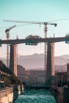 A massive bridge under construction at sunrise with cranes and mountains in the background.