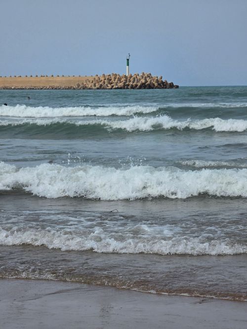 Free Serene view of ocean waves hitting the shoreline with a distant lighthouse on the breakwater. Stock Photo