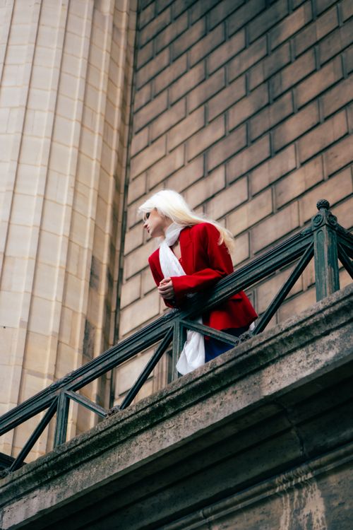 Free Stylish woman in red coat gazing from a historic balcony, capturing urban elegance. Stock Photo
