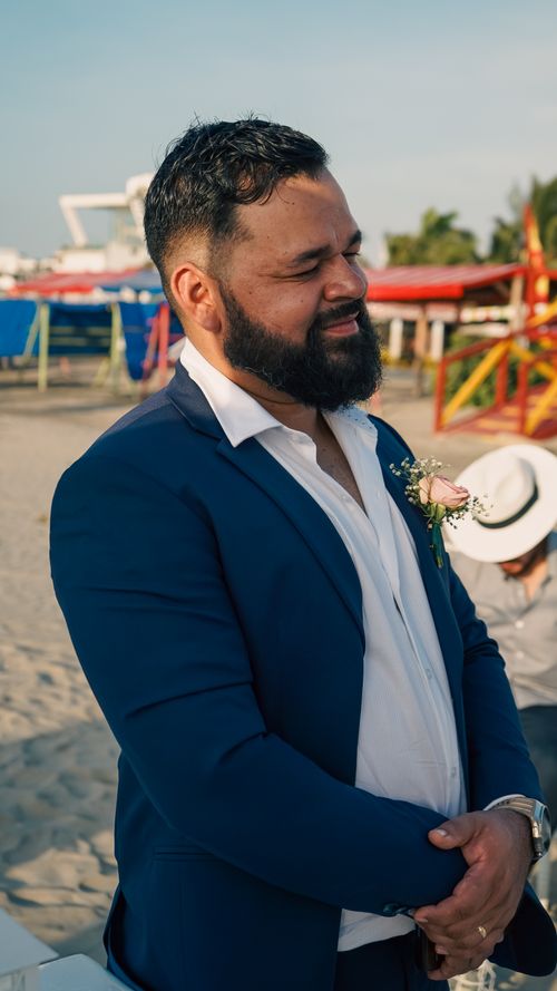 Free Smiling groom in a navy suit at a beach wedding ceremony, warmly lit by the afternoon sun. Stock Photo