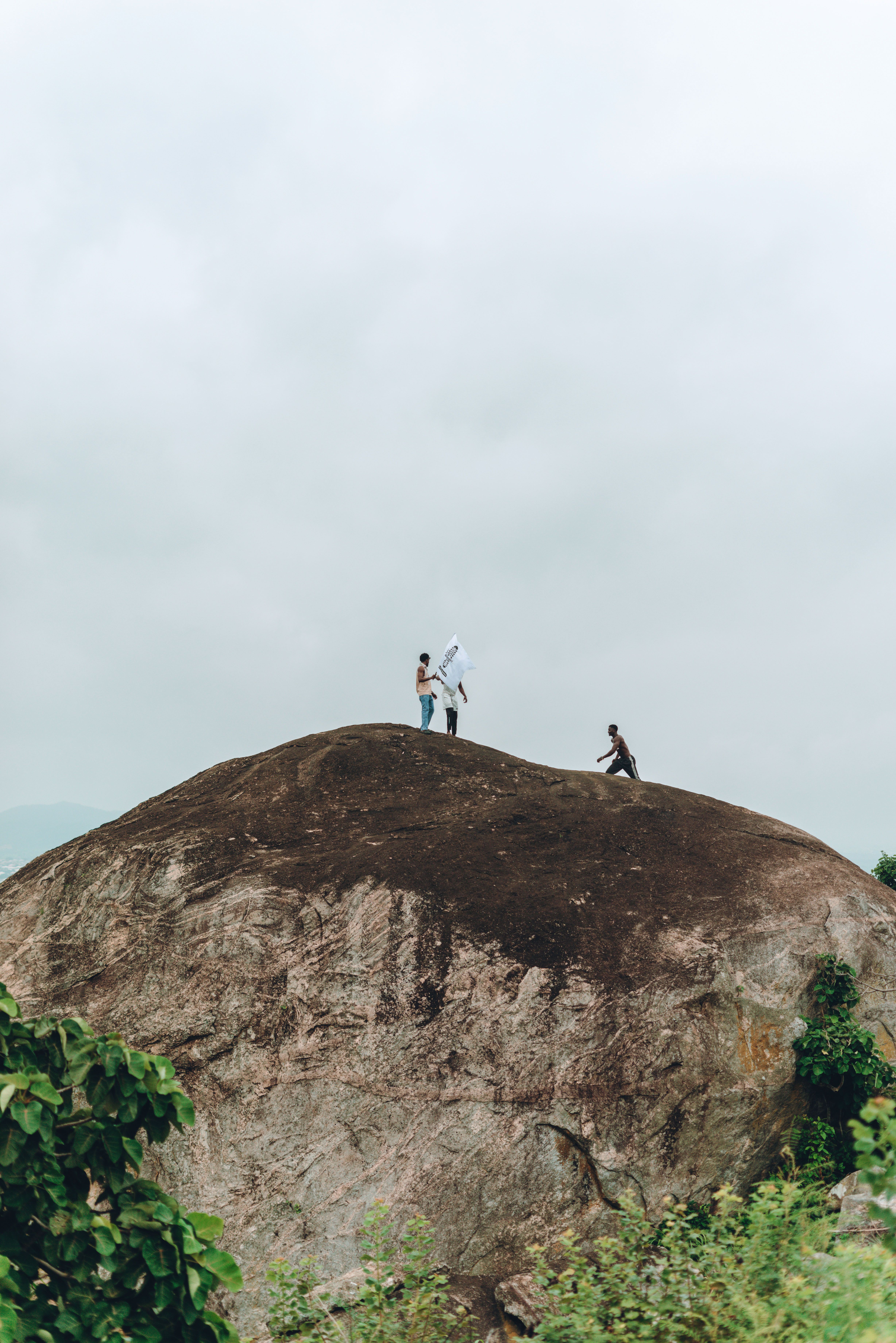 Fotomural Tres personas haciendo senderismo sobre una gran roca panorámica bajo un cielo nublado, disfrutando de la aventura.