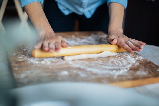 Hands rolling dough with a wooden rolling pin on a floured surface.