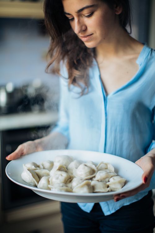 Free A young woman proudly holds a plate of freshly made dumplings in a cozy kitchen setting. Stock Photo