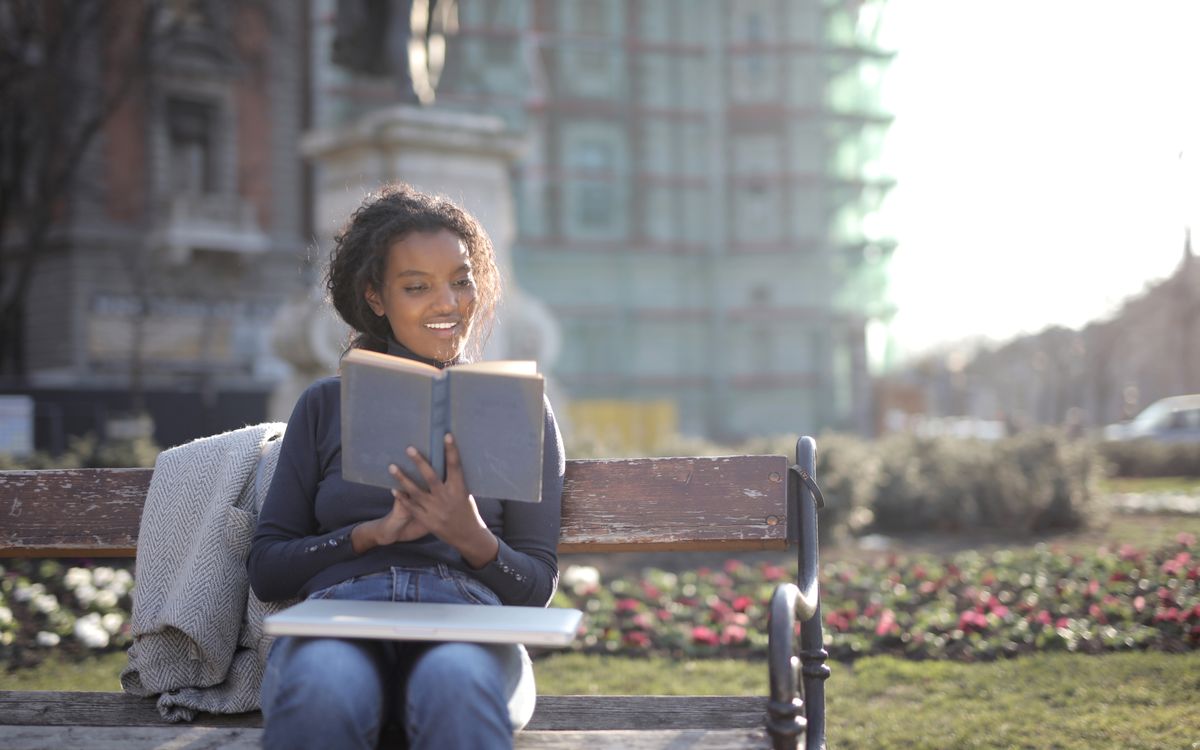 Smiling woman reading a book on a bench in a sunny park setting.