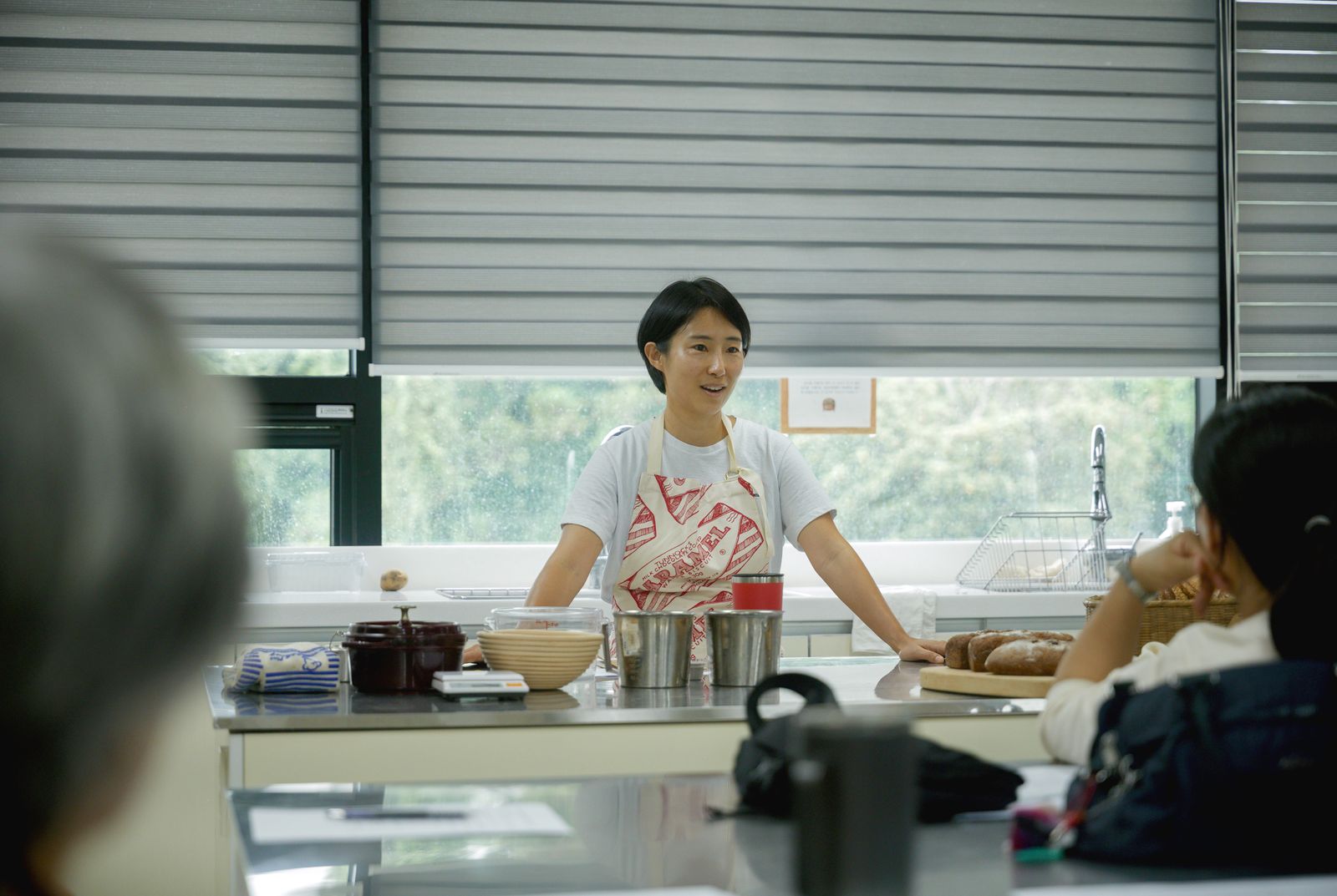 A woman in a patterned apron leading a cooking class at a steel counter, students in the foreground