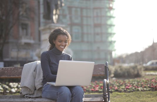 A woman enjoys working remotely on a laptop in a sunny outdoor urban park setting.