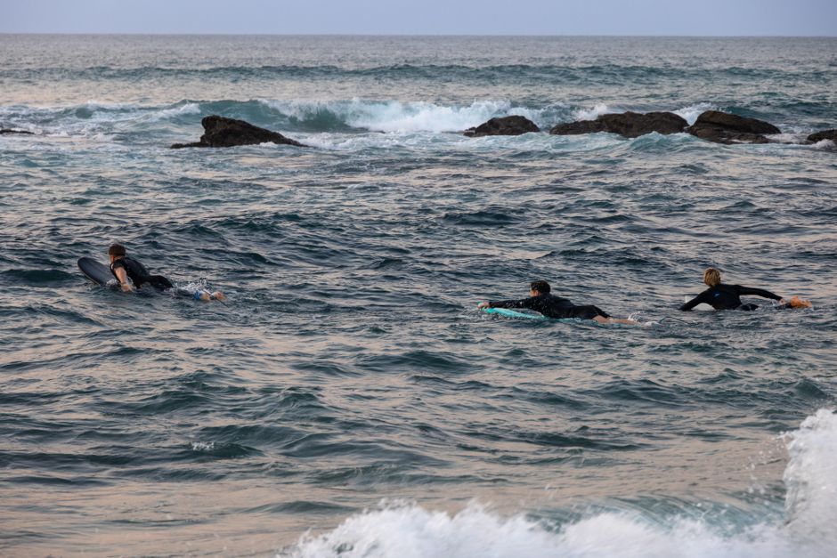 Three surfers paddle into ocean waves near rocky coast at dusk, preparing to catch waves.