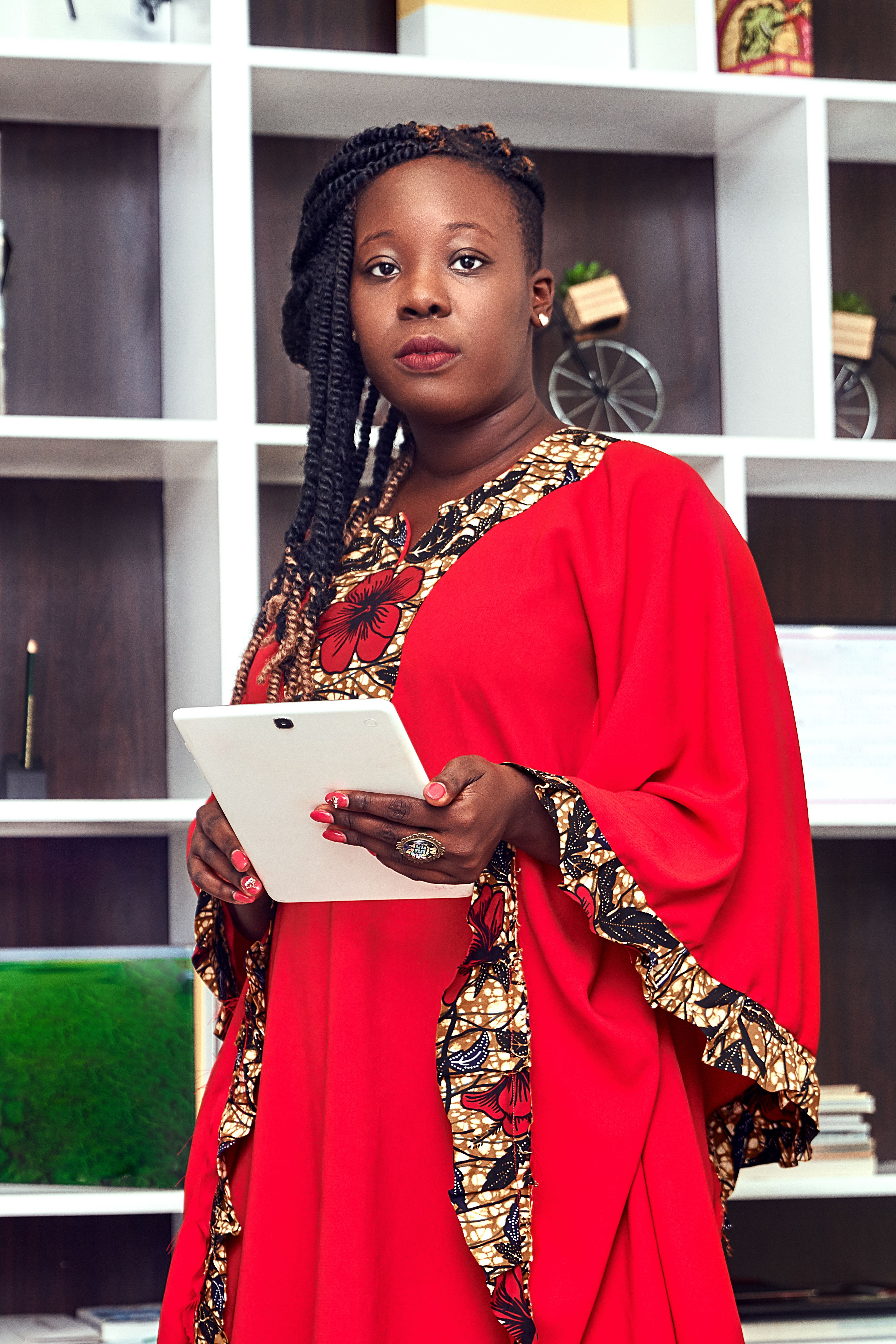 Businesswoman in a red dress holding a tablet, standing confidently in a modern office setting.