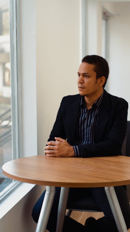 Man in Black Formal Coat Wearing Blue and Black Stripped Shirt Sitting at a Table Looking Out the window