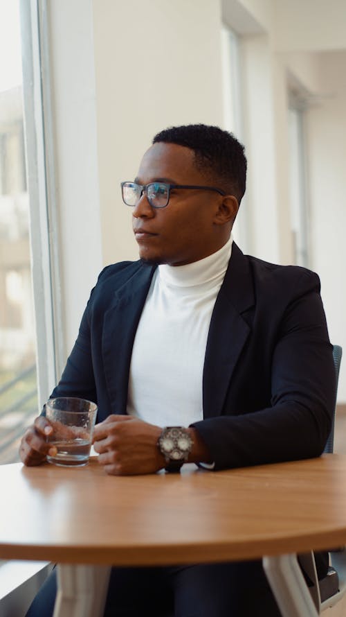 Pria Mengenakan Jas Formal Hitam Dan Kemeja Turtleneck Putih Memegang Gelas Minum Bening Melihat Keluar Jendela