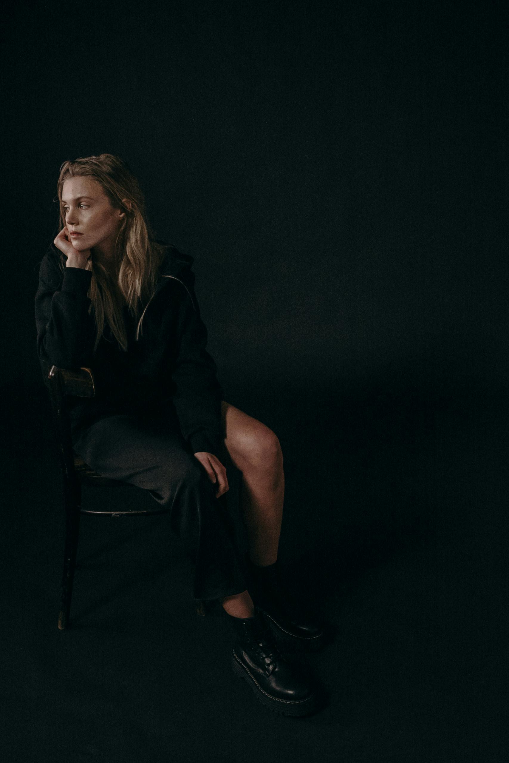 一个女人穿着深色服装，若有所思地坐在黑色背景的椅子上