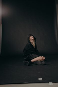 一个穿着黑色连帽衫的女人，脸上带着轻松的微笑，坐在黑暗的工作室地板上