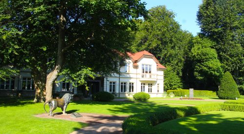 Free Charming villa exterior with lion statue surrounded by vibrant green garden and trees. Stock Photo