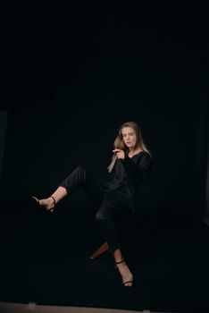 Stylish woman in black outfit posing on a stool against a dark background.