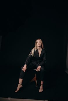 Portrait of a stylish woman in a dark fashion outfit sitting confidently on a wooden stool in a studio.