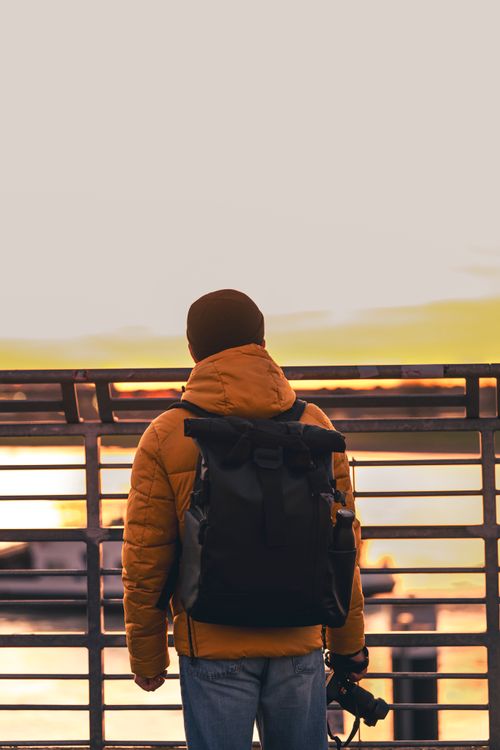 Free A man with a backpack stands by a railing, viewing a sunset over water in Düsseldorf. Stock Photo