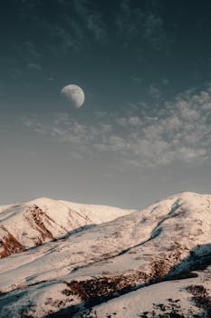 Serene snow-covered mountains under a crescent moonlit sky in winter, perfect for nature wallpapers.