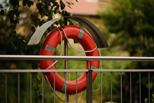 Free A bright red lifebuoy hangs on a metal railing set against a blurred green outdoor background. Stock Photo
