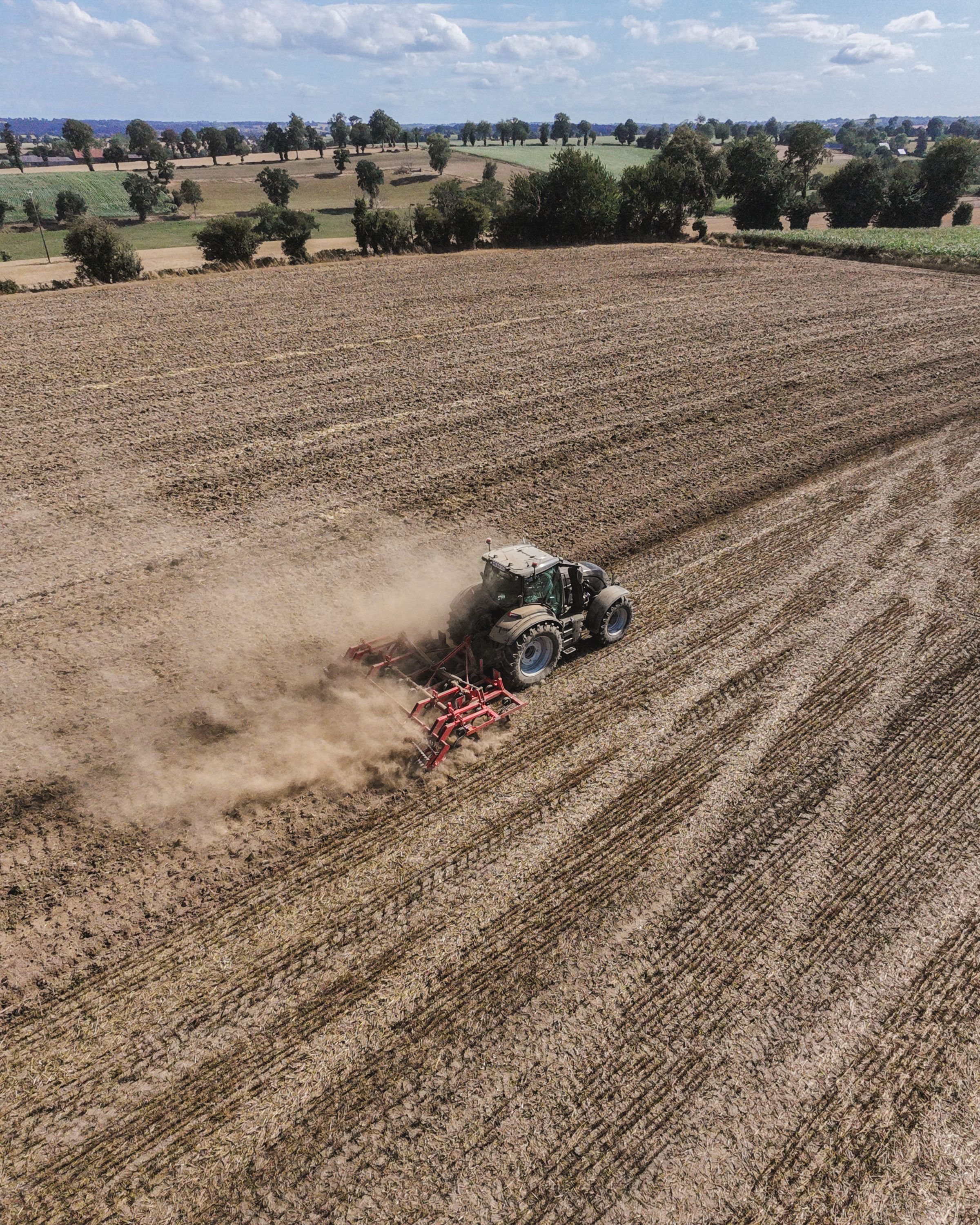 Drone shot capturing a tractor plowing a vast field, surrounded by rural scenery under a bright sky.