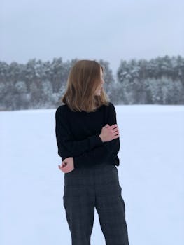 A woman stands in a snowy landscape, wearing a sweater and pants, surrounded by frosty trees.