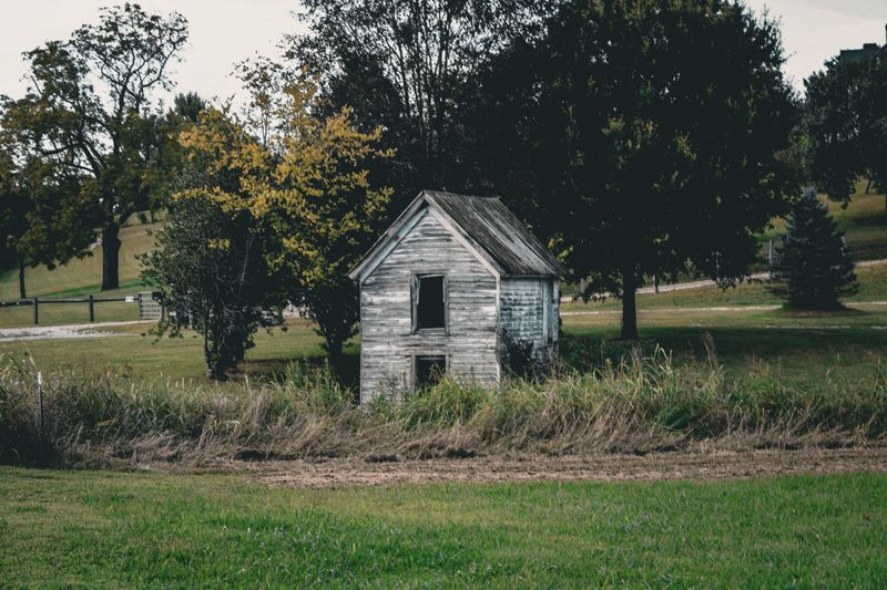 A weathered, abandoned house stands in a rural landscape amidst trees during autumn.