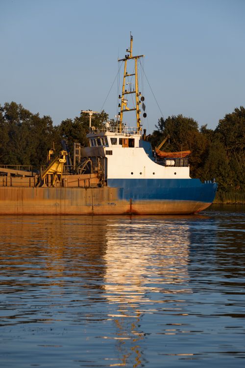 Free A large cargo ship sailing on a tranquil river during sunset, reflecting on the water. Stock Photo