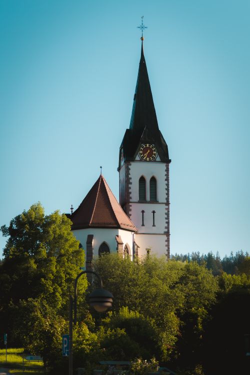Free Picturesque church with tall spire surrounded by lush greenery under a clear blue sky. Stock Photo