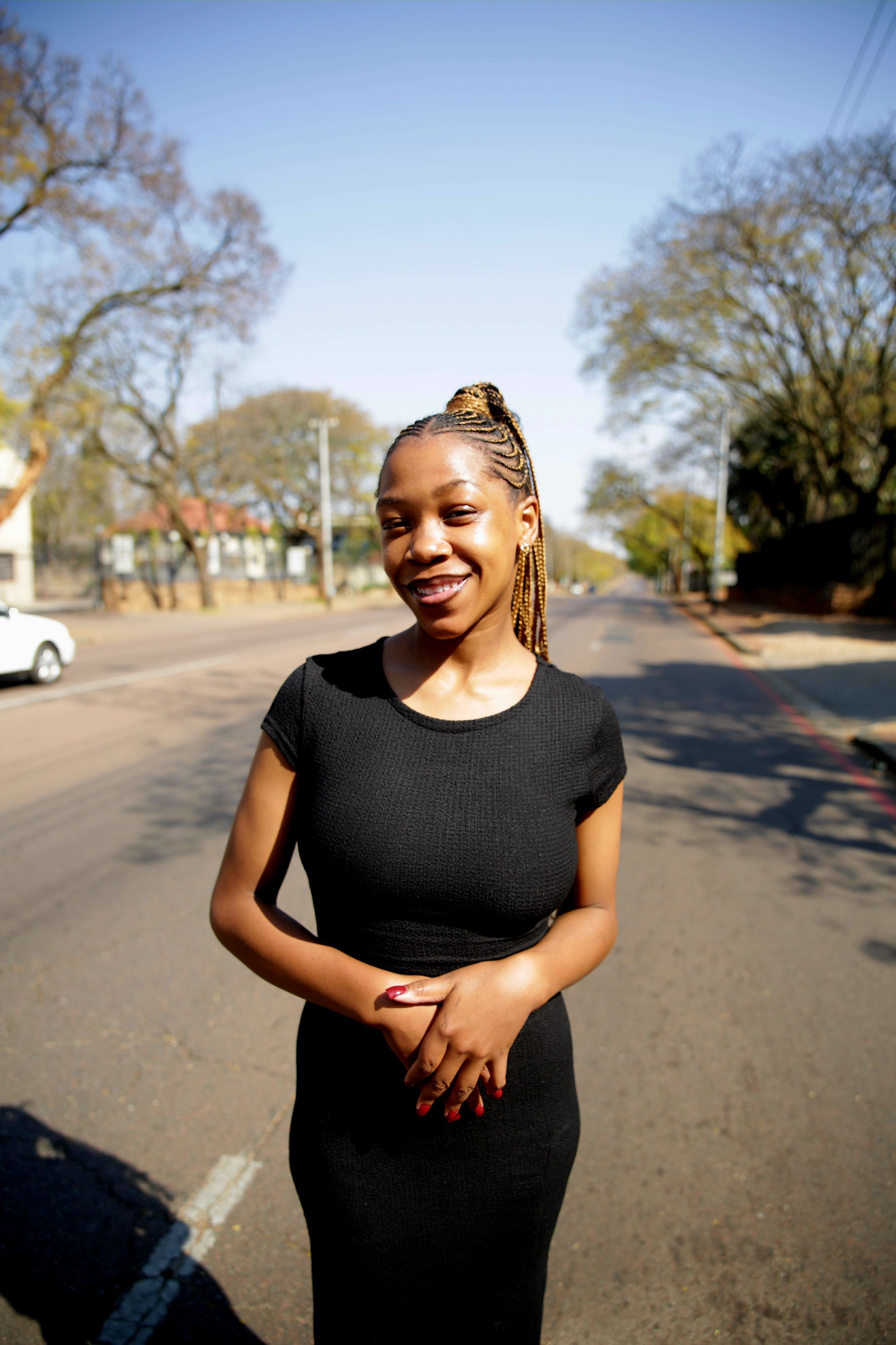 Young woman in a black dress smiling on a sunny street in Pretoria, South Africa.