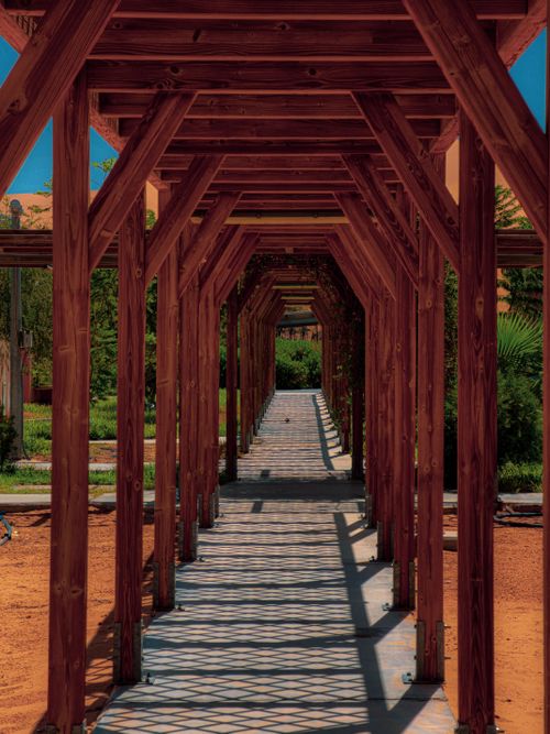 Free A long wooden walkway surrounded by lush greenery in an outdoor garden, featuring rustic architectural elements. Stock Photo