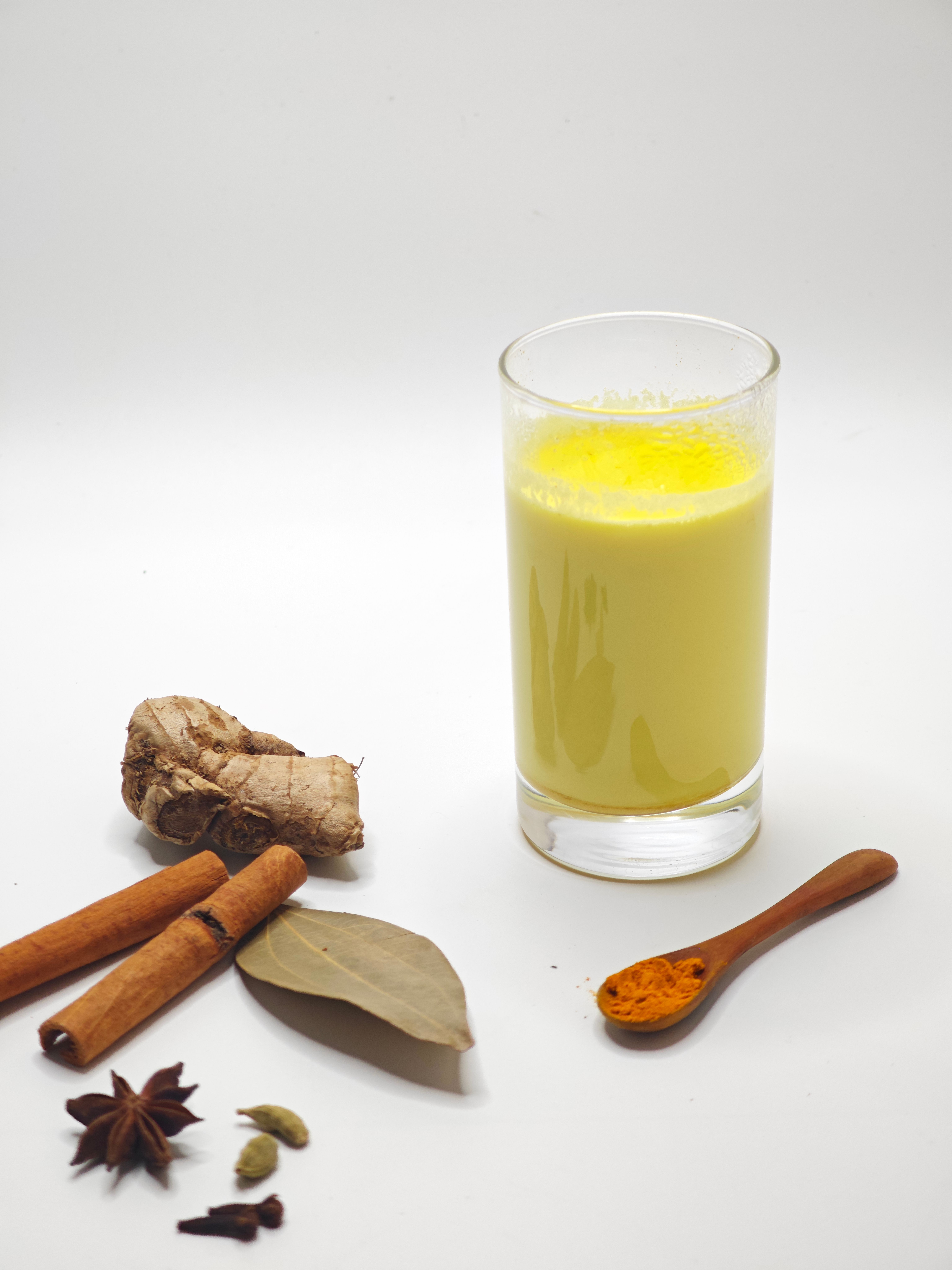 A glass of turmeric golden milk surrounded by various spices on a white background.