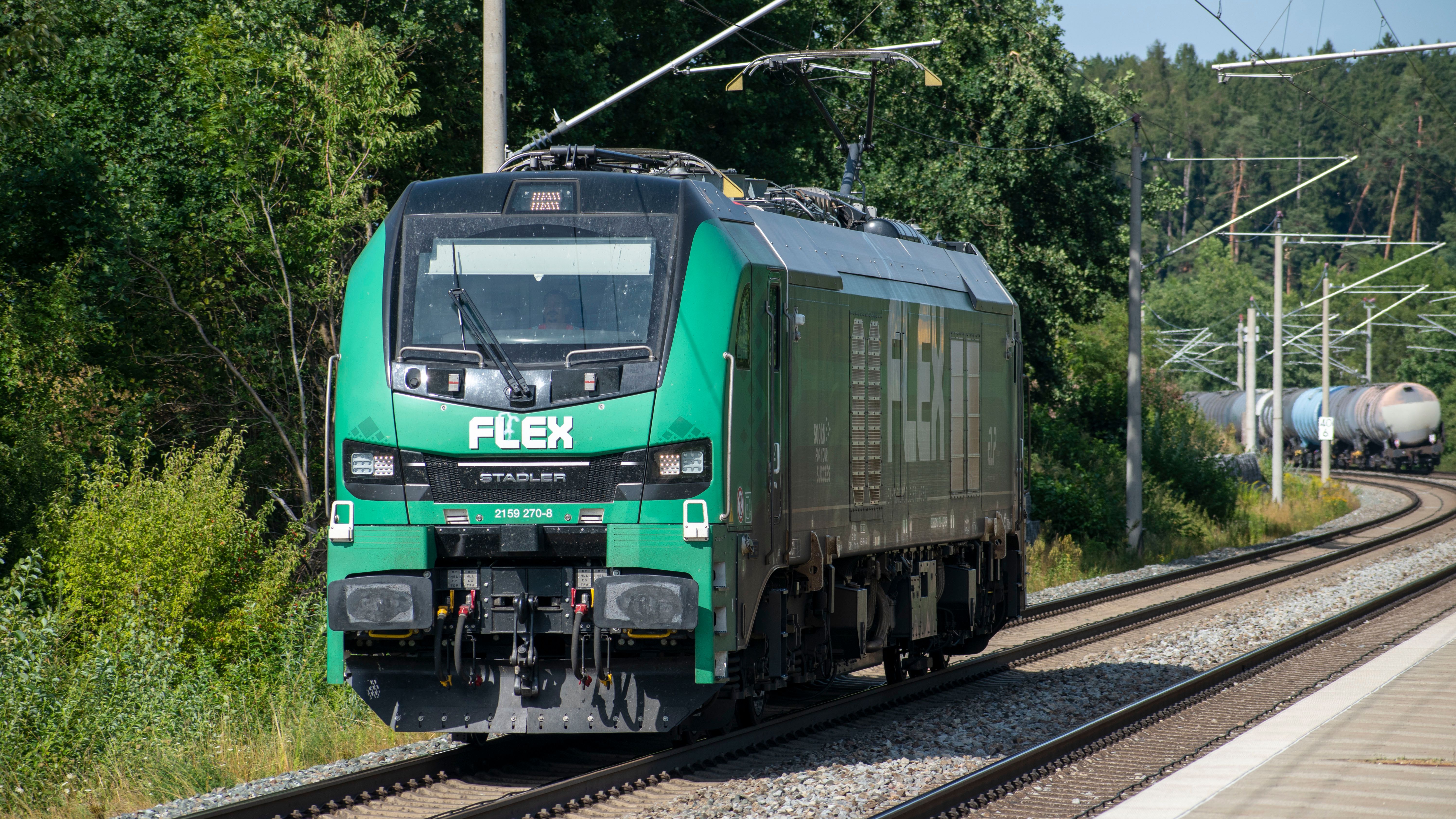A green Stadler Flex electric train travels on tracks through lush greenery in Reichertshausen, Germany.