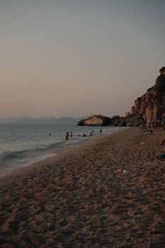A serene beach with swimmers at dusk beside rocky cliffs under a muted sky.