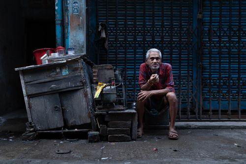 Free An elderly man in a red shirt sits alone on a sidewalk next to a wooden cart and closed shops. Stock Photo