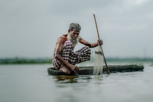 Free An elderly fisherman skillfully using a fishing net in a tranquil water body under a cloudy sky. Stock Photo