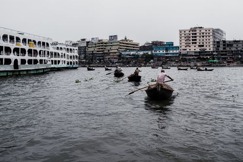 Free Boats navigate a bustling river scene near Dhaka's vibrant waterfront, highlighting urban life. Stock Photo