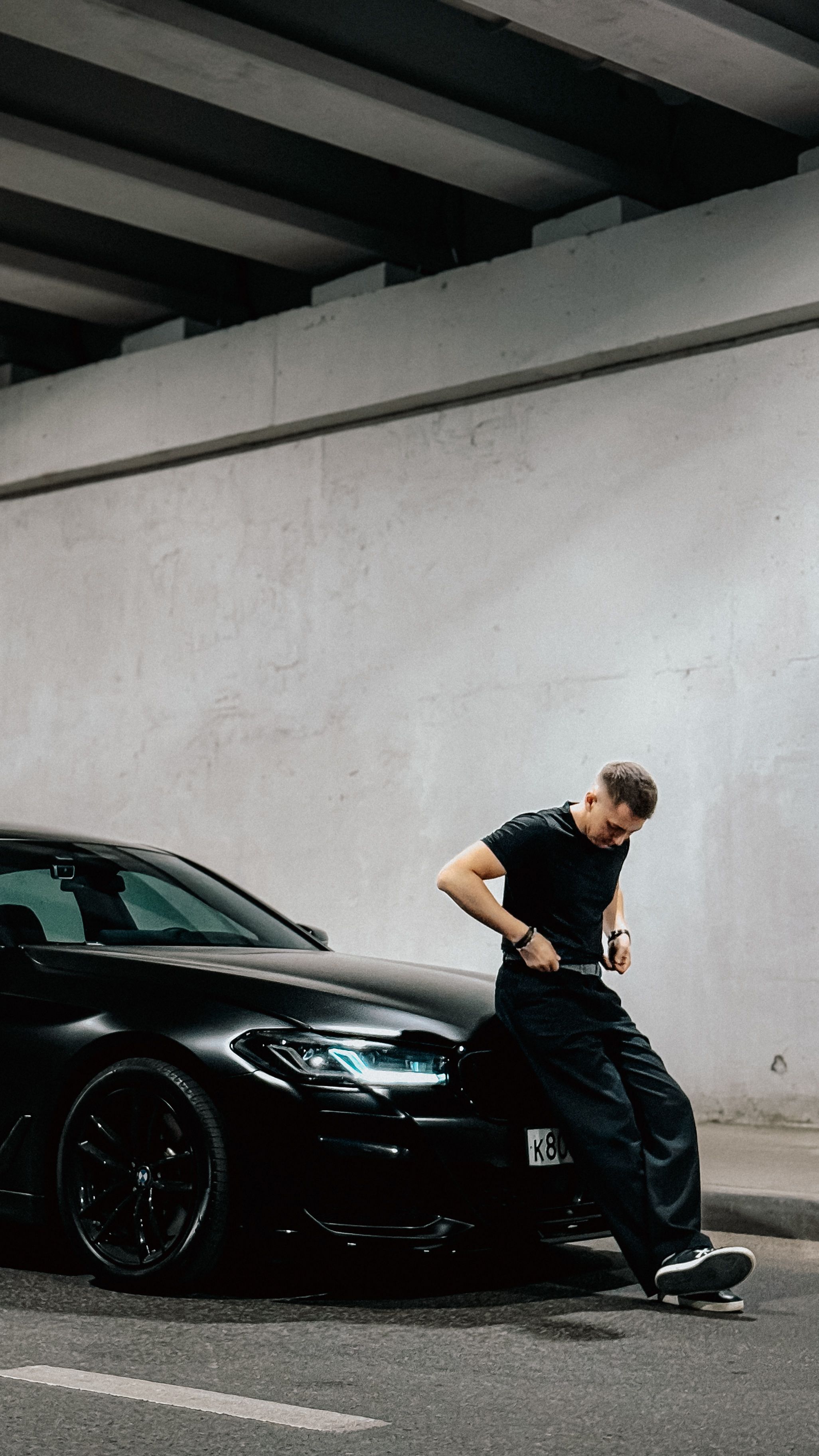 Stylish young man in black outfit casually leans on a sleek black sports car in an urban tunnel.