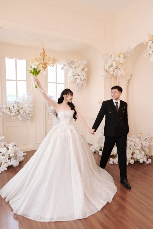Free Bride and groom in elegant wedding attire holding hands surrounded by floral decor indoors. Stock Photo