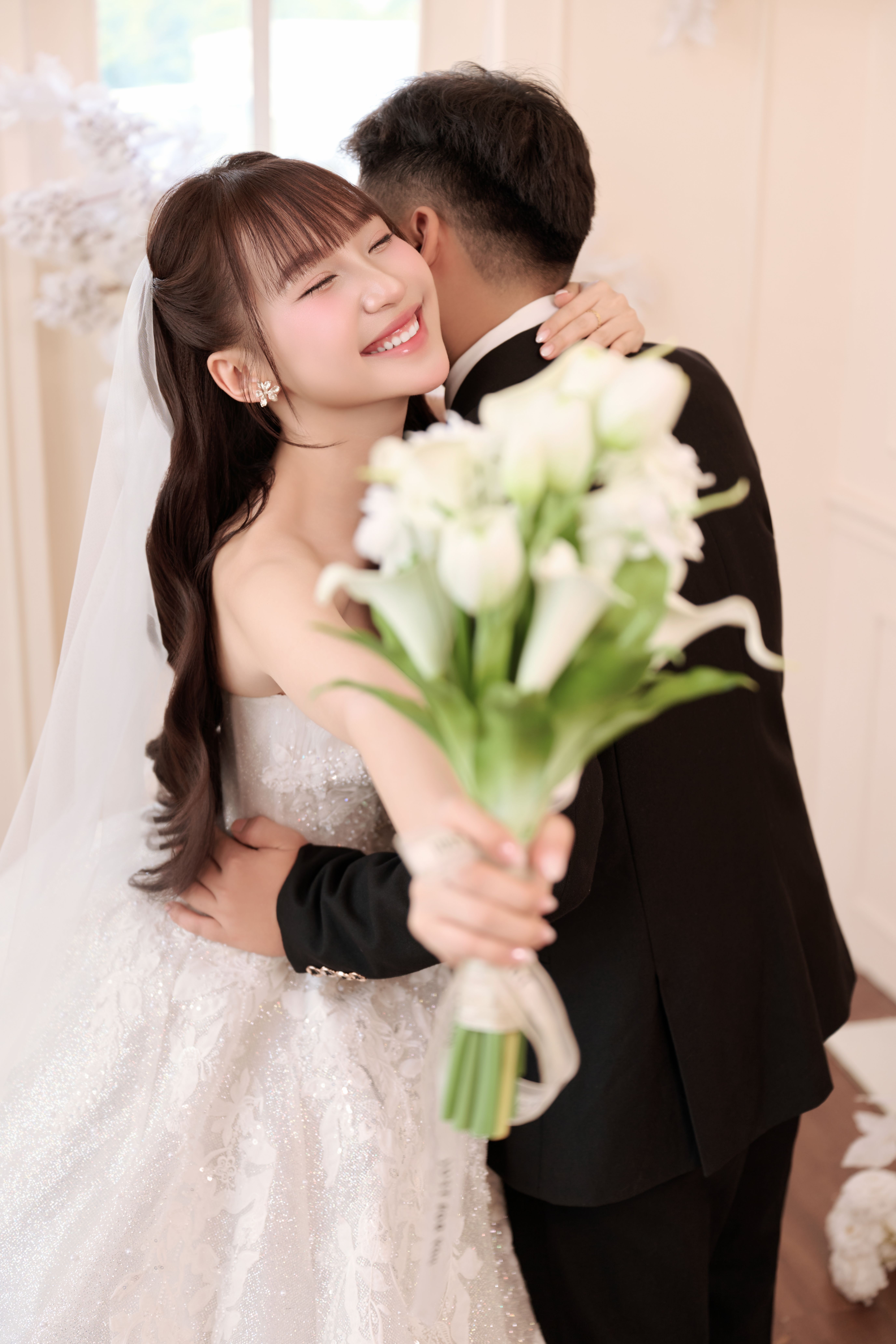 Happy bride in white dress hugs groom tightly, holding bouquet.