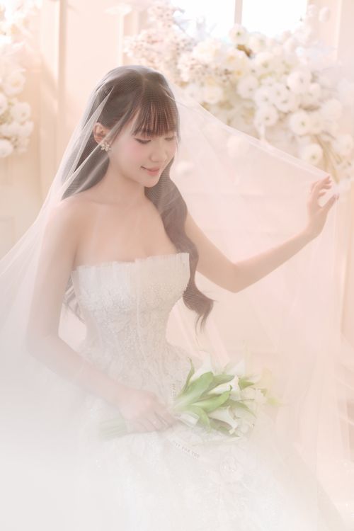 Free Beautiful bride in lace dress holding lilies, surrounded by white flowers in a soft, romantic indoor setting. Stock Photo