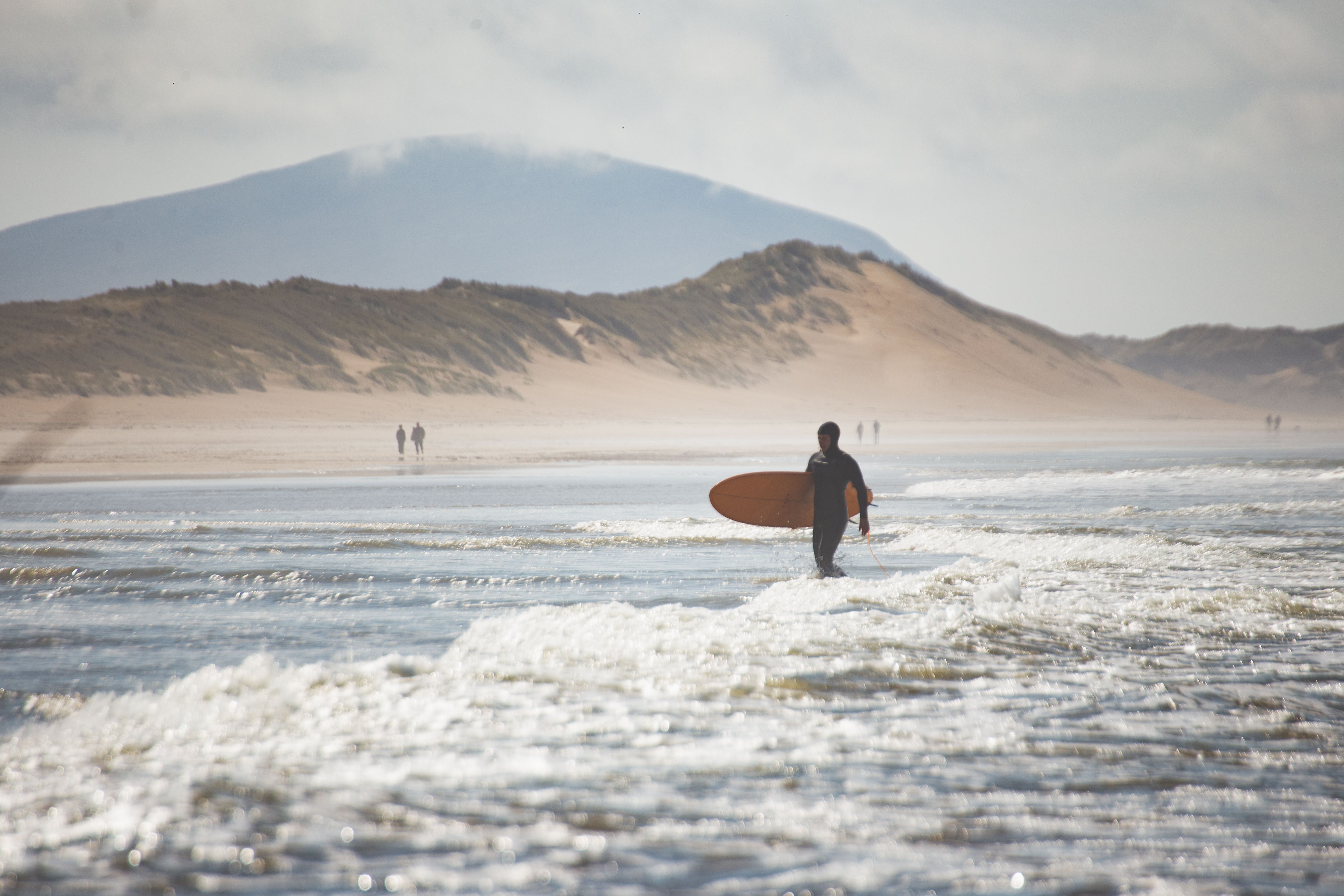 A surfer carrying a board on an Irish beach with dunes and misty hills in the background.