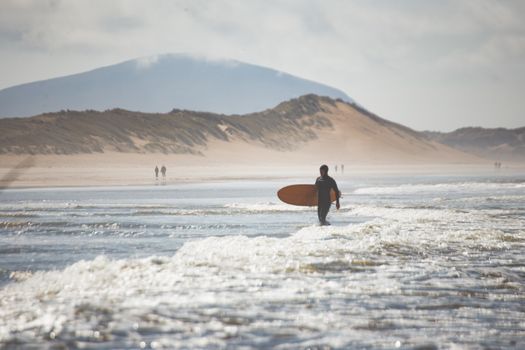 A surfer carrying a board on an Irish beach with dunes and misty hills in the background.