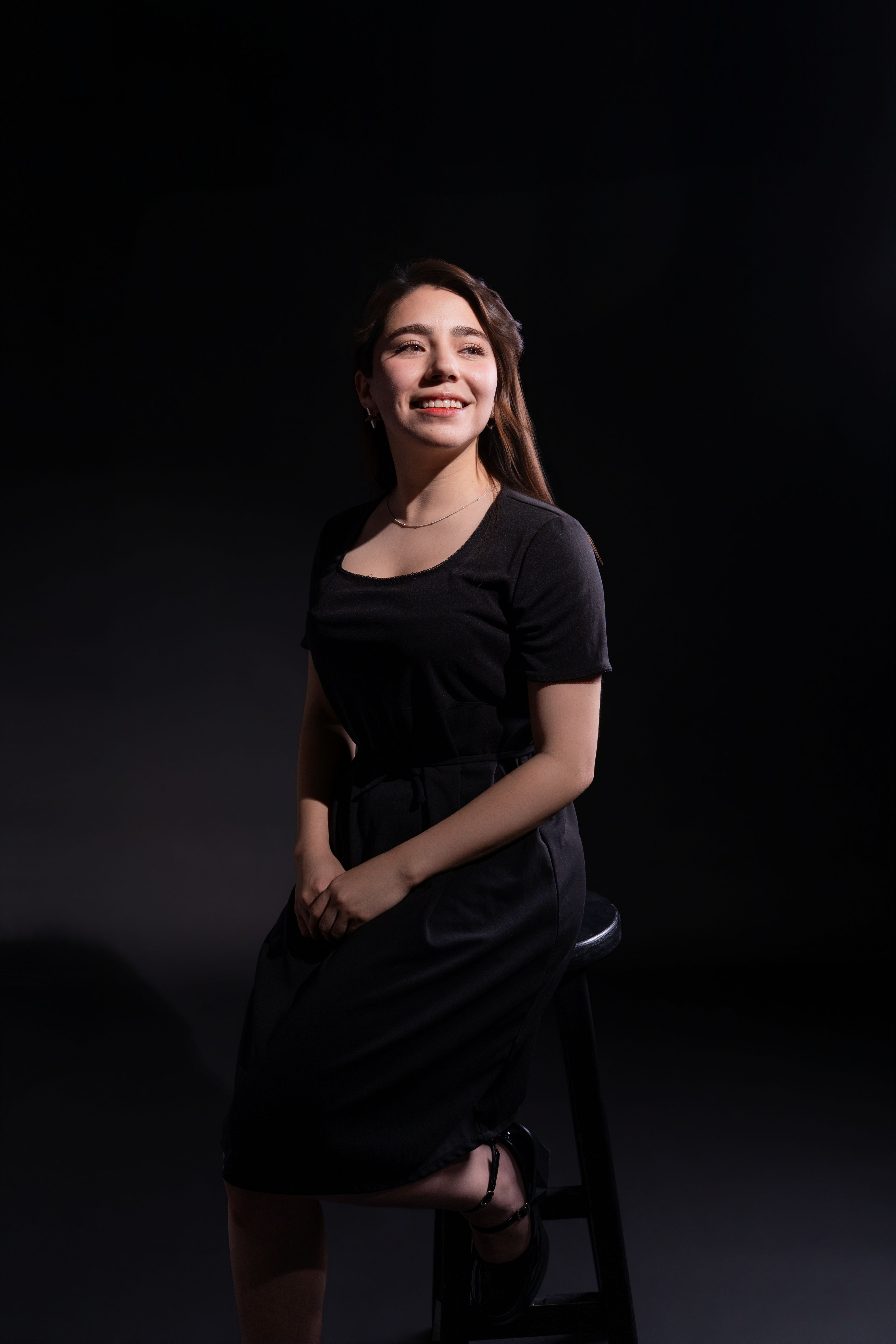 Elegant woman sitting on stool, smiling with confidence in studio lighting.