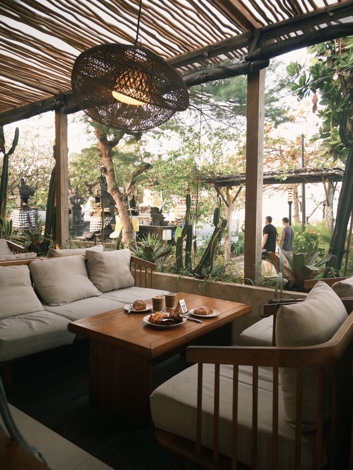A shaded lounge with low sofas, timber tables and tropical plants under a thatched roof