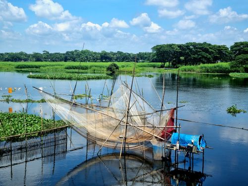 Free Traditional fishing net setup over a serene rural waterbody, surrounded by lush greenery under a bright blue sky. Stock Photo