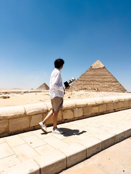 A photographer with a camera walks near the iconic Pyramids in Egypt under a clear blue sky.