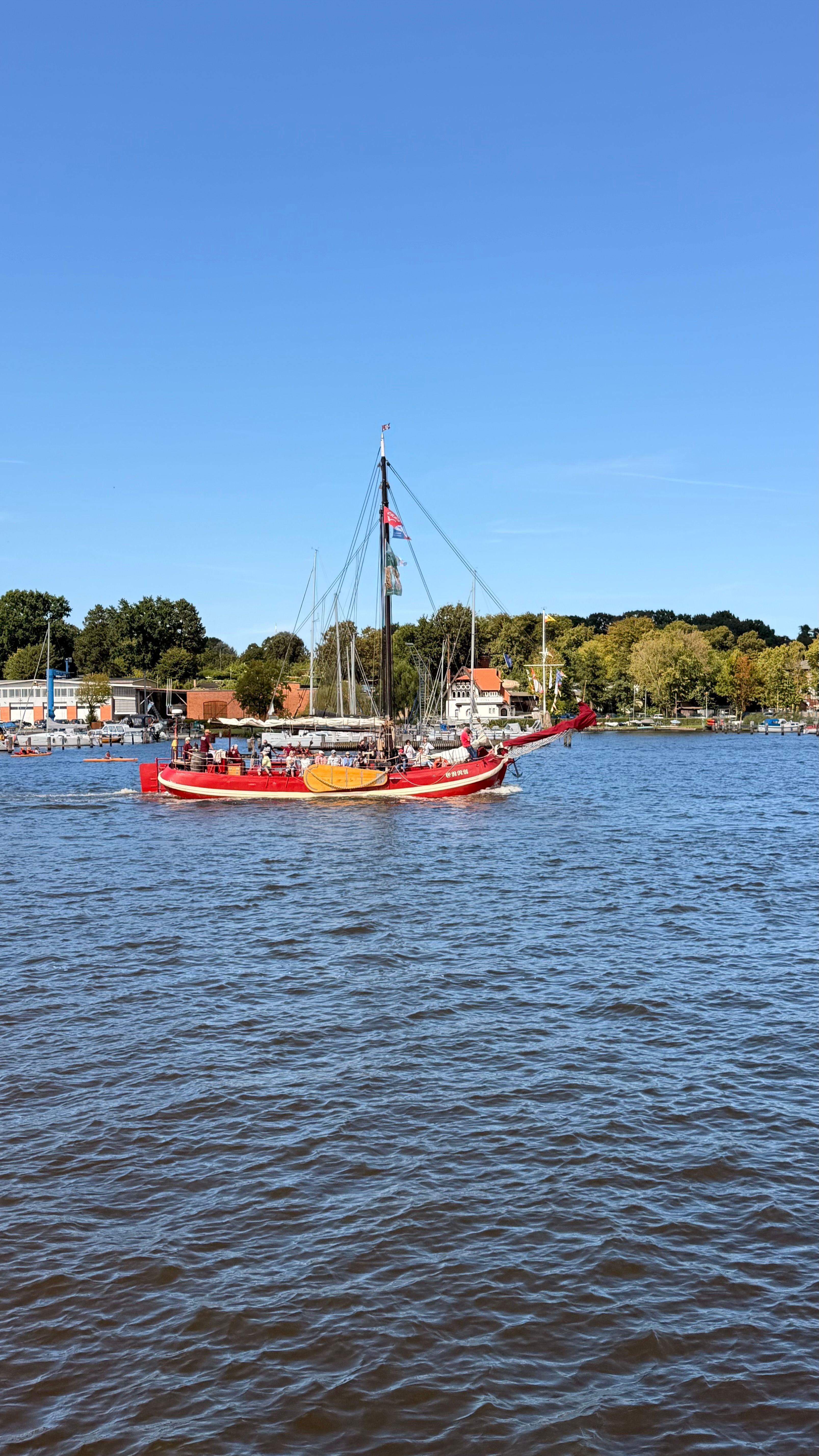 A vibrant sailboat sails along the river in the scenic city of Rostock, Germany under a clear blue sky.