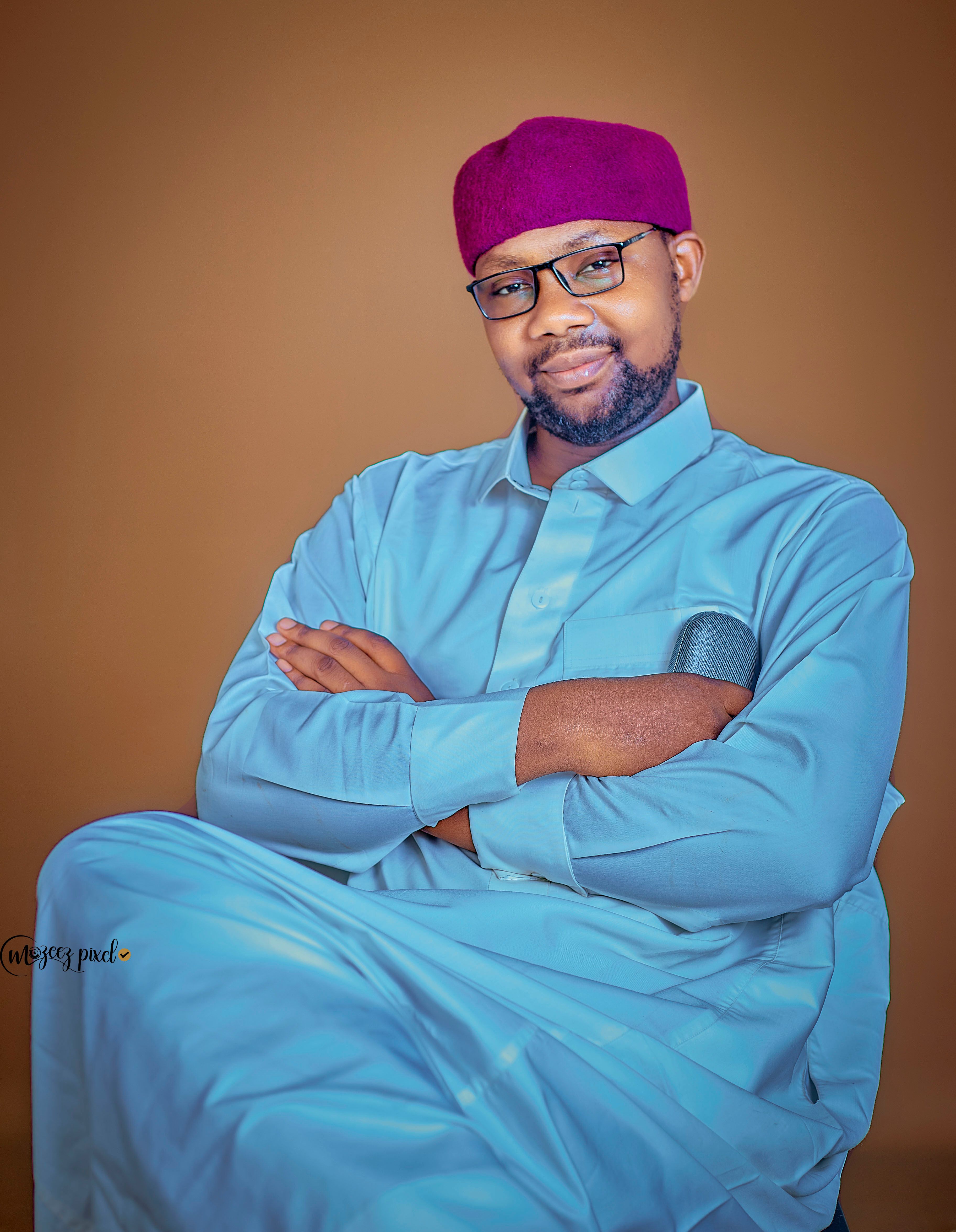 Smiling man in traditional attire with a red cap and glasses, arms crossed.