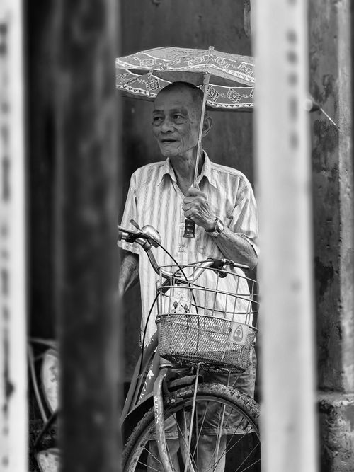 Free Elderly man stands with his bicycle under an umbrella in a Kolkata street, captured in black and white. Stock Photo